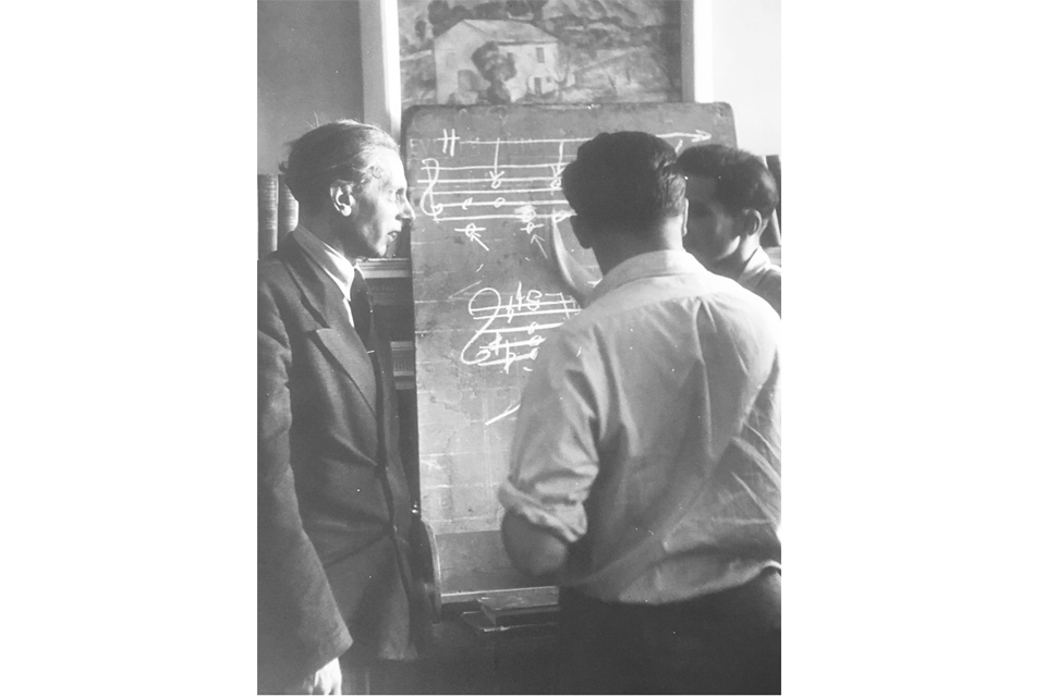 An old photograph of two men, wearing suits and shirts, looking at a black board with chalk music notes.